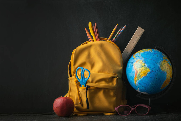 Yellow backpack with school supplies next to the globe, red apple and glasses on the black school board background. Back to school concept on September day - Photo, image