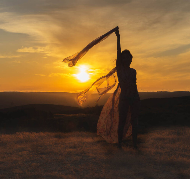 силуэт женщины на закате с тканью в руках и прозрачным платьем, которое движется ветер - Фото, изображение