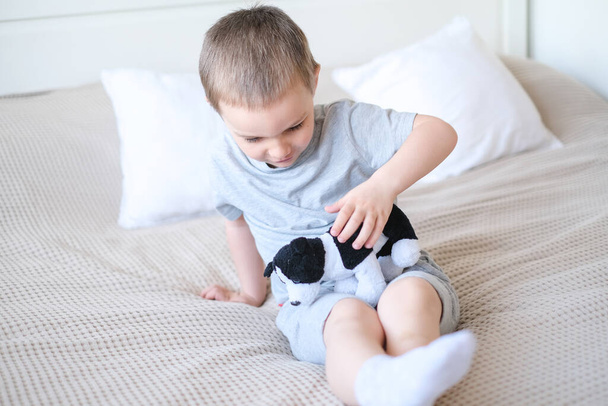 Close-up of a small happy caucasian boy in a gray t-shirt lies on the bed with a toy dog. - Valokuva, kuva