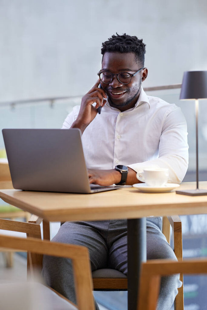 Африканский бизнесмен многозадачный, сидя в кафе. Бизнесмен печатает на ноутбуке и разговаривает по телефону с клиентом. - Фото, изображение