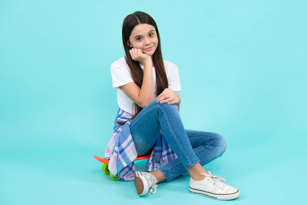 Fashion girl wearing trendy t-shirt and jeans sit on skateboard, posing on blue background. Fashion teenager, casual look. Happy teenager, positive and smiling emotions of teen girl - Foto, Imagem