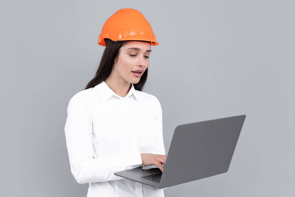 Young woman in helmet hold laptop computer on gray background. Architect businesswoman in helmet thinking, young caucasian female in work as constructor engineer - Fotó, kép