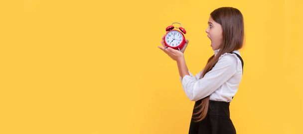 shocked teen girl in school uniform with alarm clock showing time on yellow background, punctuality. Teenager child with clock alarm, horizontal poster. Banner header, copy space - Fotografie, Obrázek