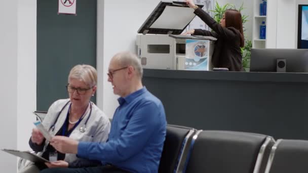 Medical worker sitting at facility reception desk to help people with healthcare appointments and patients in waiting room. Receptionist using checkup visit reports at counter in lobby. - Video