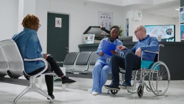 Senior patient in wheelchair doing consultation with nurse in waiting area lobby, examining man with chronic impairment and disability. Specialist and person at checkup visit. Tripod shot. - Filmagem, Vídeo