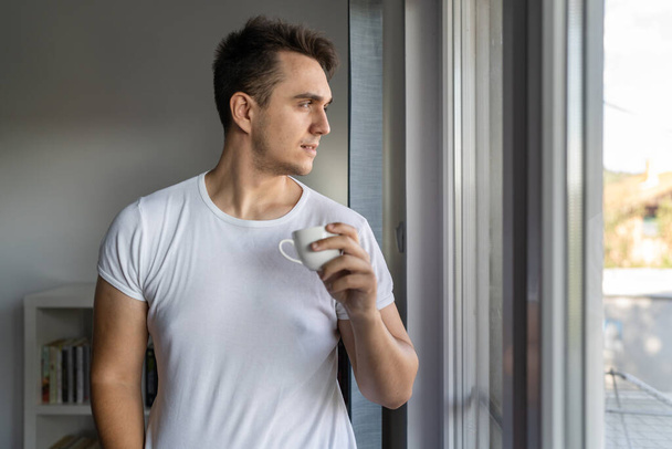 One man young adult male caucasian with brown hair standing by the window at home in morning day confident looking to the side thinking contemplation wear white shirt with cup of coffee copy space - Photo, Image