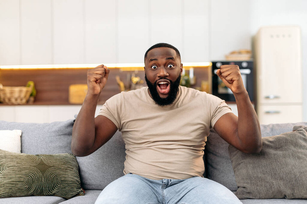 Ενθουσιασμένος χαρούμενος γοητευτικός νεαρός Αφροαμερικάνος, κάθεται στον καναπέ στο σαλόνι, ενθουσιασμένος για την επιτυχία με τα χέρια σηκωμένα, γιορτάζοντας τη νίκη χαμογελώντας ευτυχισμένος, ουρλιάζοντας. Νικήτρια ιδέα - Φωτογραφία, εικόνα