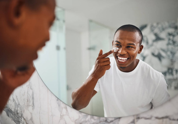 Taking his skincare routine to the next level. a handsome young man applying moisturizer to his face in the bathroom at home - Foto, imagen