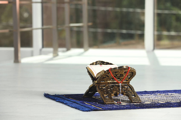 Rehal with open Quran and Misbaha on Muslim prayer rug near window indoors, space for text - Photo, Image