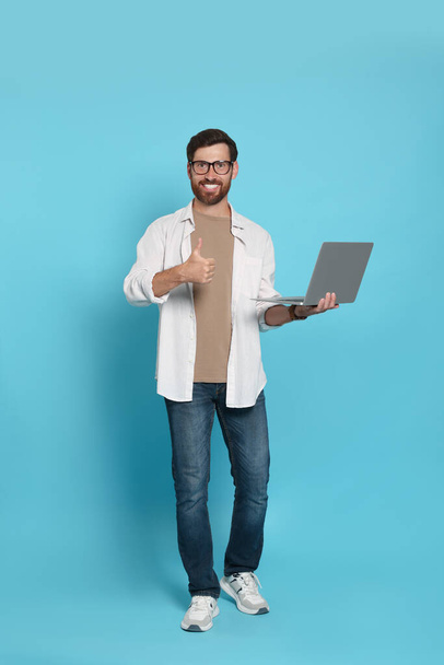 Handsome man with laptop showing thumb up gesture on light blue background - Photo, Image