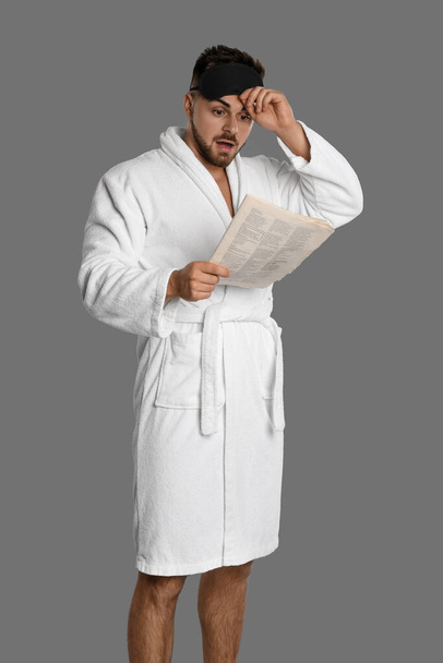 Young man in bathrobe reading newspaper on grey background - Foto, Imagen
