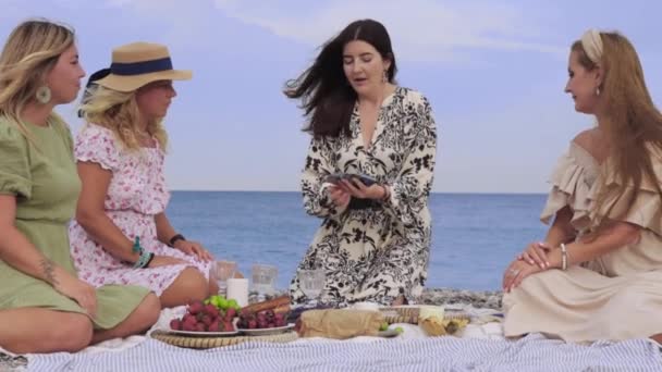 Four adult women at a picnic by the sea playing a card game. Mid shot - Materiał filmowy, wideo