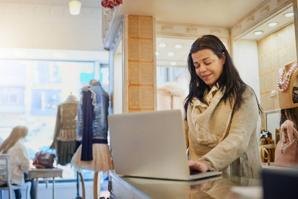 Using online resources to manage her business. an attractive mature female entrepreneur working on a laptop in her self-owned boutique - Photo, Image