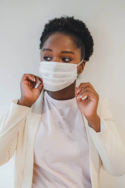 Black female with curly hair putting on medical mask while standing in a room with closed air, and looking away. Isolated over white wall. Respiratory virus - Фото, зображення