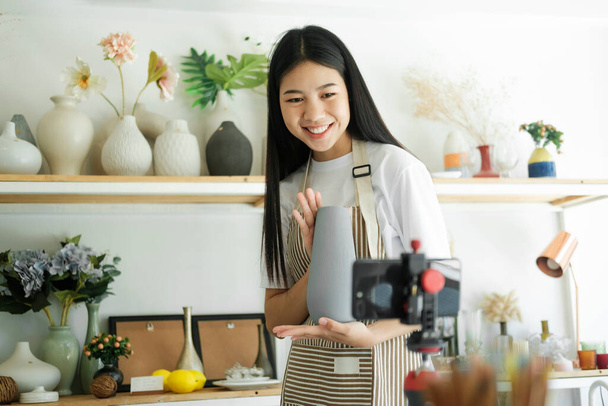 Young asian businesswoman fashion blogger showing ceramic products with smiling face while recording new content for vlog with camera at home studio. - Foto, Imagen