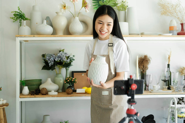 Young asian businesswoman fashion blogger showing ceramic products with smiling face while recording new content for vlog with camera at home studio. - Foto, imagen