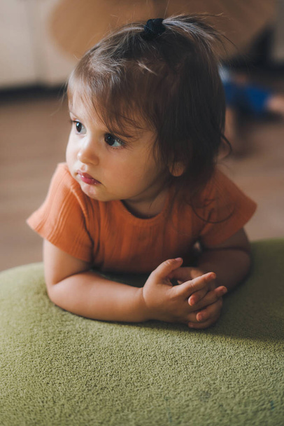 Baby girl pretty face with beautiful deep eyes looking away while sitting on the floor leaning on green sofa. Relax and peace. Innocent infant expression - 写真・画像