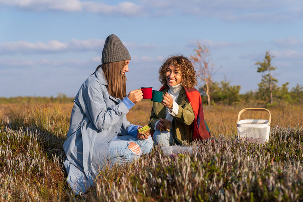 Happy girl best friends drink hot beverage in bright mugs sitting at picnic against blue sky. Women enjoy to spend autumn weekend uniting with nature and discuss issues sitting on grass meadow closeup - Photo, Image