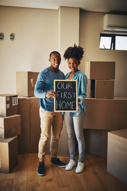 This is where our life begins. Portrait of a cheerful young couple holding up a sign saying our first home together while being surrounded by cardboard boxes inside at home - Fotografie, Obrázek