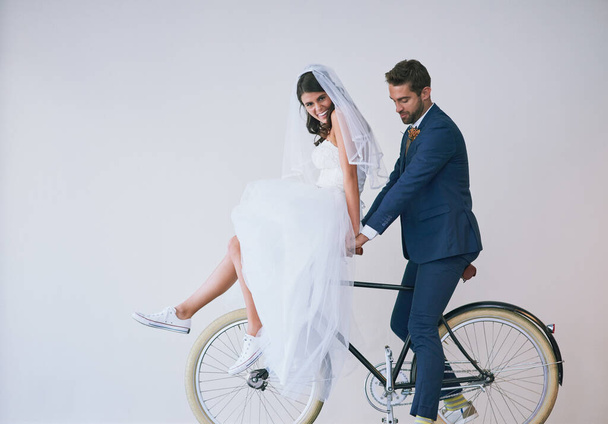 Marriage, one awesome ride. Studio shot of a newly married young couple riding a bicycle together against a gray background - Photo, Image