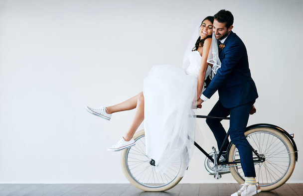 Off to our honeymoon. Studio shot of a newly married young couple riding a bicycle together against a gray background - Photo, image