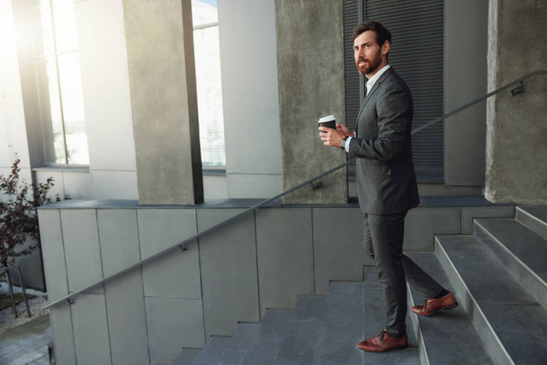Όμορφος επιχειρηματίας κατεβαίνει τις σκάλες με καφέ κατά τη διάρκεια του διαλείμματος κοντά στο γραφείο. Θολή φόντο - Φωτογραφία, εικόνα