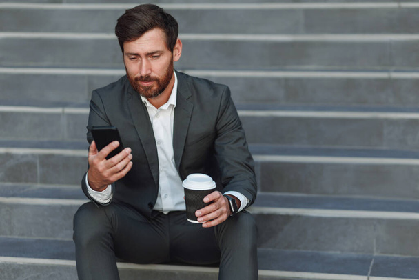 Бизнесмен в костюме с телефоном пьет кофе возле офисного здания. Концепция бизнеса - Фото, изображение