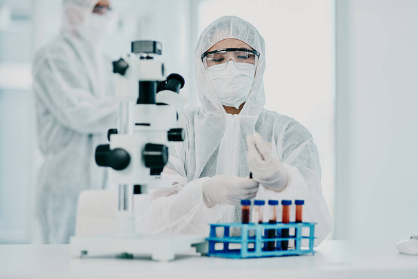 A scientist conducting a medical experiment to research covid vaccine in a lab and collecting blood samples. Healthcare researcher analyzing chemicals in a chemistry laboratory wearing a hazmat suit. - Foto, Bild