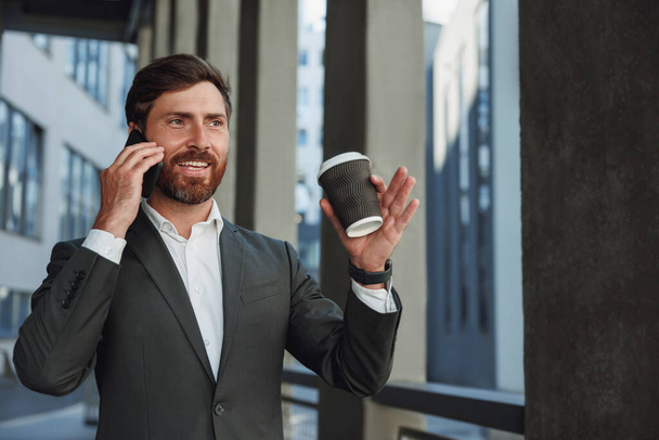 Smiling businessman in suit with coffee talking phone near office building. Business concept - Photo, Image