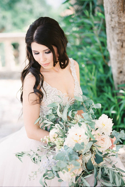 Bride with a bouquet of flowers sits with her head bowed under a tree. High quality photo - Foto, Imagem