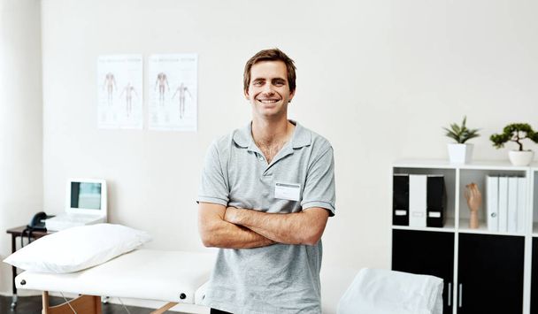 We put our clients first. Portrait of a confident young male physiotherapist standing with his arms folded inside of his office - Фото, зображення