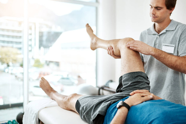 Держите его крепче. молодой физиотерапевт-мужчина помогает клиенту с упражнениями для ног, кто лежит на кровати - Фото, изображение