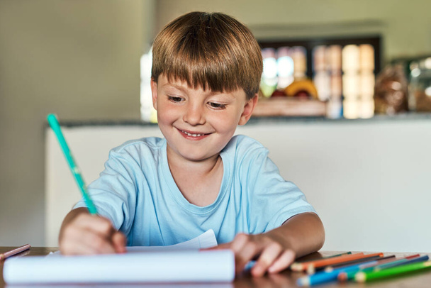 Η ώρα της εργασίας είναι χαρούμενη. ένα μικρό αγόρι κάθεται σε ένα γραφείο και γράφει σε ένα σημειωματάριο στο σπίτι - Φωτογραφία, εικόνα