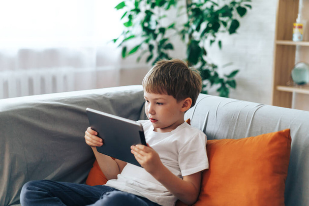 Мальчик сидит на диване и смотрит видео на планшете.. - Фото, изображение