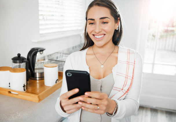 Γυναίκα που χαζεύει, στέλνει μηνύματα και διαβάζει σε ένα τηλέφωνο στην κουζίνα της στο σπίτι. Χαμογελώντας γυναίκα στα μέσα κοινωνικής δικτύωσης σε απευθείας σύνδεση app, δικτύωση και μηνύματα επαφές, ενώ χαμογελώντας σε ένα αστείο post, meme ή βίντεο. - Φωτογραφία, εικόνα