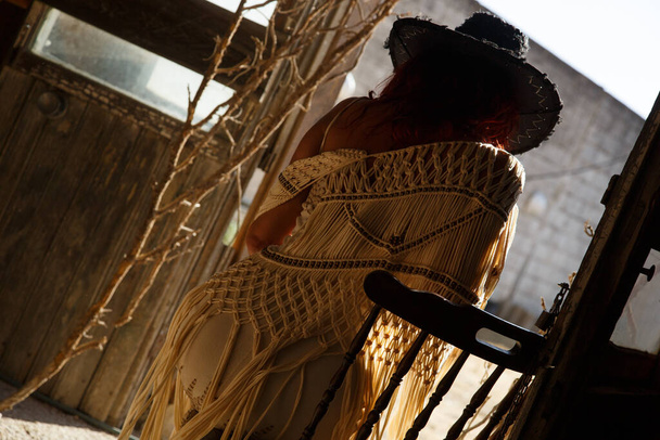 Γυναίκα οπίσθιο φως στην πόρτα κάθεται σε καρέκλα με καπέλο στο παλιό βιομηχανικό κτίριο - Φωτογραφία, εικόνα