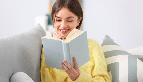 Beautiful young woman reading interesting book on sofa at home - Photo, image