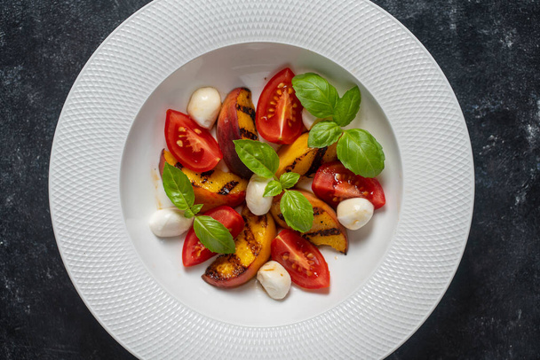 Grilled peach salad with mozzarella, tomato, green basil and sauce. Fresh gourmet salad in white plate on black background, close up, top view - Photo, Image