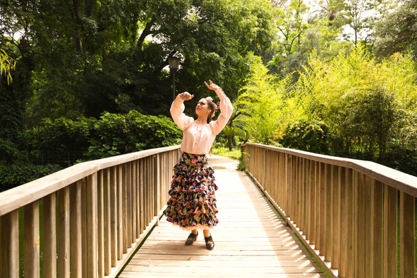 Beautiful teenage flamenco dancer dressed in a flowery skirt with ruffles and flowers in her hair is dancing in the park on a wooden bridge. Flamenco concept cultural heritage of humanity. - Fotoğraf, Görsel