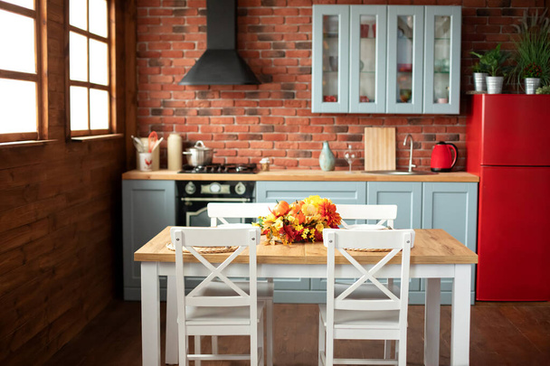 Κομψή τραπεζαρία με ξύλινο τραπέζι και καρέκλες. Ζεστή κουζίνα διακοσμημένη με φθινοπωρινή διακόσμηση και τραπεζομάντηλα. Σκανδιναβική κουζίνα εσωτερικού σχεδιασμού με σκεύη, πιάτα, πιάτα.  - Φωτογραφία, εικόνα