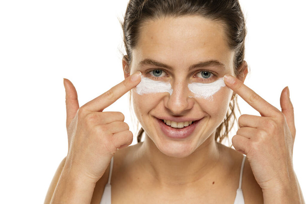 portrait of a young smiling woman applying cosmetic product under her eyes on a white background. - Photo, Image