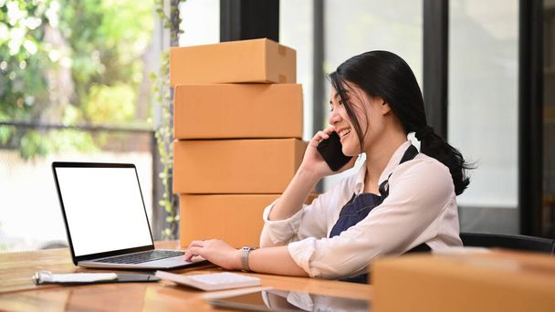 Small business entrepreneur having phone conversation and confirming orders from customer on laptop. Online selling concept. - 写真・画像