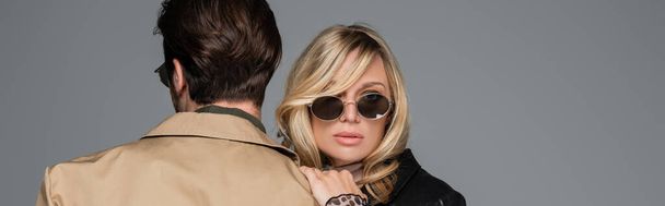blonde in stylish sunglasses hugging man in beige jacket isolated on grey, banner - Photo, Image