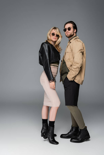 full length of stylish woman in autumnal outfit and sunglasses posing near man standing with hand in pocket on grey - Foto, Imagem