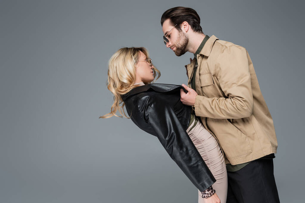 side view of bearded man in sunglasses pulling leather jacket of blonde woman while flirting isolated on grey - Photo, Image