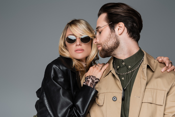 blonde and stylish woman in sunglasses hugging bearded man isolated on grey - Foto, afbeelding
