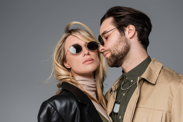 stylish bearded man in sunglasses looking at blonde woman in black leather jacket isolated on grey - Foto, Imagen