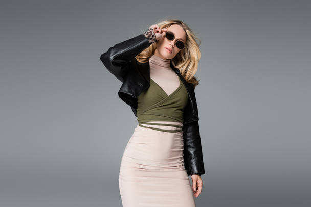 blonde woman in black leather jacket adjusting trendy sunglasses isolated on grey - Photo, Image