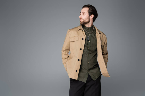 bearded man in trendy outfit posing with hands in pockets and looking away isolated on grey - Foto, Imagem