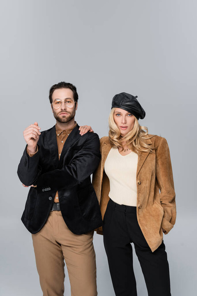blonde woman in stylish beret posing with hand in pocket near bearded man in sunglasses isolated on grey - Zdjęcie, obraz